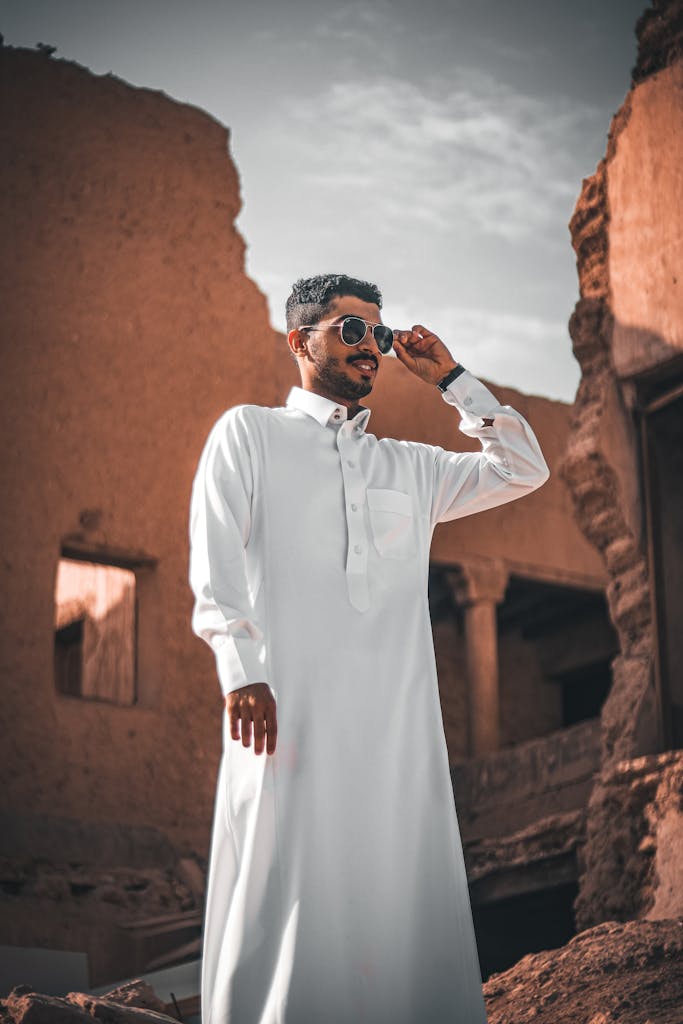 A fashionable man in traditional attire stands confidently among ancient ruins in Riyadh, Saudi Arabia.